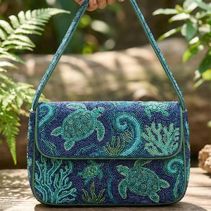 Puede incluir: Un bolso de mano con cuentas con un tema marino. El bolso azul marino presenta tortugas marinas y corales en tonos de verde y azul. El bolso tiene un cierre de solapa y una correa verde. El bolso está sostenido por una mano.