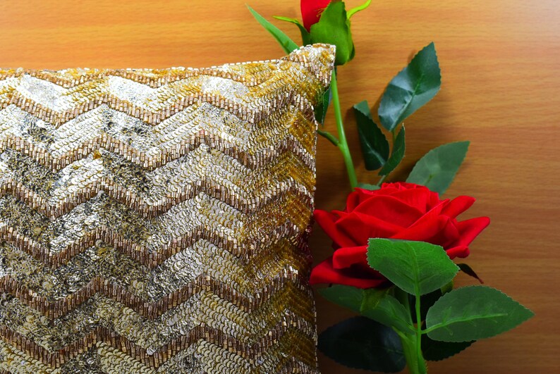K&ouml;nnte beinhalten: Ein Kissen mit goldenen und silbernen Pailletten in einem Chevron-Muster, neben einer roten Rose mit gr&uuml;nen Bl&auml;ttern.