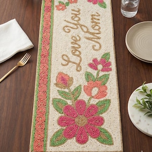 Puede incluir: Camino de mesa con cuentas con el texto "Love You Mom" en letras doradas. El camino de mesa presenta un diseño floral con flores y detalles de cuentas rosas, naranjas y verdes. El borde es una mezcla de cuentas rosas y verdes.