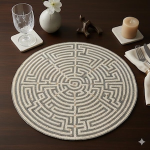 May include: A round beaded placemat with a maze design in gray and off-white. The placemat is on a dark brown wooden table, with a glass, coaster, candle, and vase in the background.