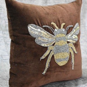 May include: A brown velvet pillow with a beaded bee design. The bee is made of silver and gold beads and has a detailed design.