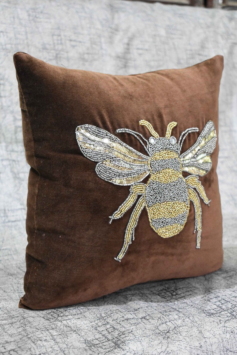 May include: A brown velvet pillow with a beaded bee design. The bee is made of silver and gold beads and has a detailed design.