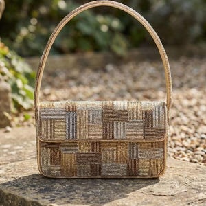 Puede incluir: Bolso de mano rectangular con cuentas y asa curva. El bolso presenta un diseño de patchwork en tonos dorados, marrones y plateados. El asa y el ribete son dorados. El bolso está sobre una superficie de piedra.