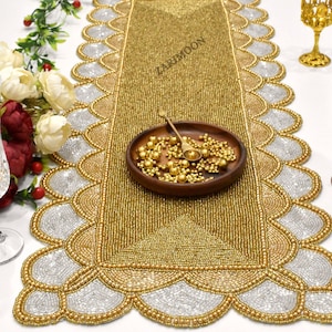 May include: Gold beaded table runner with a scalloped edge and a silver beaded design. The runner is on a white table with a glass of champagne and a red glass.