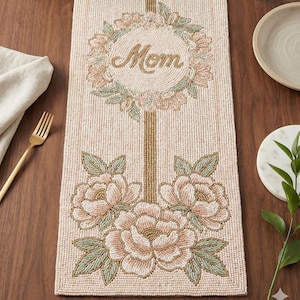 Puede incluir: Camino de mesa con cuentas con un diseño floral. El camino de mesa es beige con una corona floral y la palabra "Mom" en el centro. Debajo, tres grandes flores rosas con hojas verdes. Un tenedor y una servilleta dorados a la izquierda.