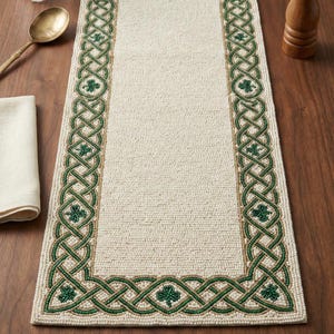 May include: Cream-colored table runner with a green Celtic knot design and shamrock accents. The border features a detailed, interwoven pattern. The runner is made of a textured material, perfect for adding a touch of Irish heritage to your dining table.