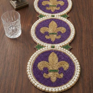 May include: A beaded table runner featuring three connected circular designs. Each circle has a purple center with a gold fleur-de-lis, surrounded by white beads. Green accents connect the circles.