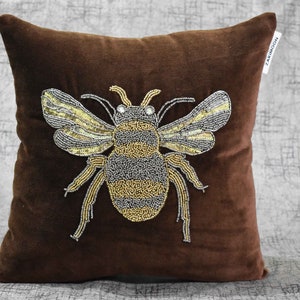 May include: A brown velvet pillow with a beaded bee design. The bee is made of gold and silver beads and has a detailed design.