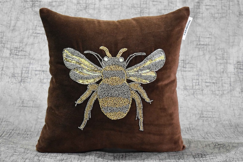 May include: A brown velvet pillow with a beaded bee design. The bee is made of gold and silver beads and has a detailed design.