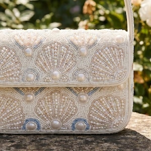 May include: A rectangular handbag featuring an intricate exterior of white, cream, and pale blue glass beads arranged in a repeating shell pattern. The design is accented with smooth, round white faux pearls integrated into the beaded surface.
