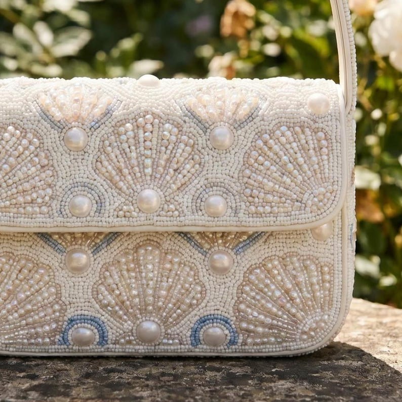 May include: A rectangular handbag featuring an intricate exterior of white, cream, and pale blue glass beads arranged in a repeating shell pattern. The design is accented with smooth, round white faux pearls integrated into the beaded surface.