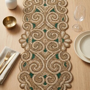 May include: Beaded table runner with an ornate design in gold, cream, and green. The runner features a scrolling pattern and is placed on a wooden table, alongside tableware and decorative items.