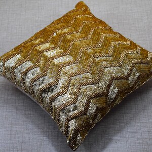 K&ouml;nnte beinhalten: Ein quadratisches Kissen mit goldenen und silbernen Pailletten und einem geometrischen Muster. Das Kissen ist weich und pl&uuml;schig.