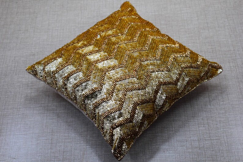 K&ouml;nnte beinhalten: Ein quadratisches Kissen mit goldenen und silbernen Pailletten und einem geometrischen Muster. Das Kissen ist weich und pl&uuml;schig.