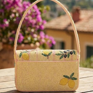 K&ouml;nnte beinhalten: Eine Perlenhandtasche mit gelbem Hintergrund und hellrosa Besatz. Die Tasche hat Zitronen- und Olivenzweig-Designs. Der Griff ist hellrosa und mit Perlen besetzt. Die Tasche steht auf einem Holztisch.