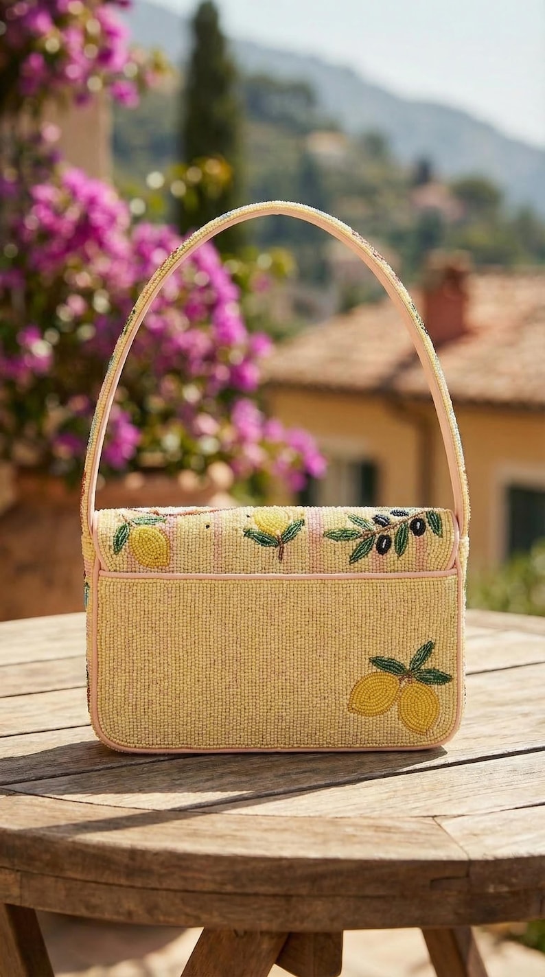 K&ouml;nnte beinhalten: Eine Perlenhandtasche mit gelbem Hintergrund und hellrosa Besatz. Die Tasche hat Zitronen- und Olivenzweig-Designs. Der Griff ist hellrosa und mit Perlen besetzt. Die Tasche steht auf einem Holztisch.