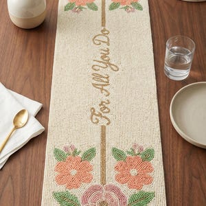 May include: Cream-coloured beaded table runner featuring floral patterns in pink, peach, and green. Gold embroidered text reads "For All You Do". The runner is rectangular.