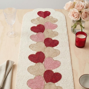 May include: Cream-colored beaded table runner with a repeating pattern of red, pink, and tan hearts. The runner is rectangular with a scalloped top edge. The hearts are arranged in a vertical line down the center of the runner. A decorative piece for Valentine's Day.
