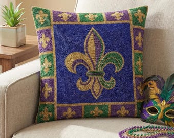 May include: A square decorative pillow with a beaded fleur-de-lis design in gold, green, and purple on a blue background. The pillow has a beaded border in green, purple, and gold. The pillow is on a beige couch.
