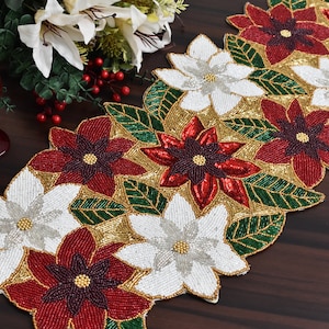 May include: A beaded table runner with a Christmas theme. The runner features white, red, and gold poinsettia flowers with green leaves.