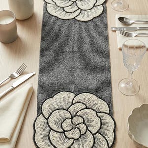 May include: A grey beaded table runner featuring large, white flower designs at each end. The flowers have black outlines and sit on a grey, textured background. Table setting with cutlery, glasses, and a candle.