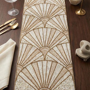 May include: A cream and gold beaded table runner with a fan design. The rectangular runner features a repeating pattern of stylized fans. The beads create a textured surface, adding visual interest to the dining table setting.
