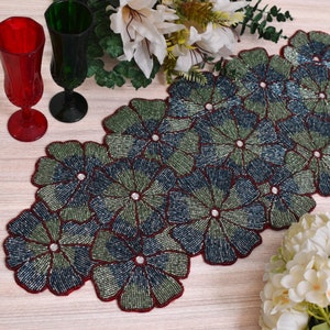 May include: A beaded table runner with a floral design in shades of green and blue. The runner has a red border and is made of small, round beads.