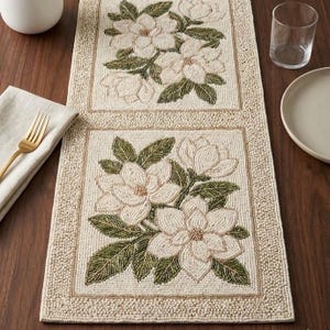 Pode incluir: Caminho de mesa com contas com um design de flores de magnólia. O caminho de mesa tem um fundo creme com flores brancas bordadas e folhas verdes. A borda é bege claro e tem formato retangular.