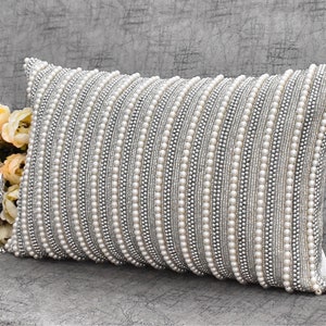 May include: A white and silver decorative pillow with a beaded design. The pillow is rectangular and has a subtle sheen. The brand name "ZAKIMOON" is printed on the back of the pillow.