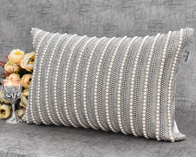 May include: A white and silver decorative pillow with a beaded design. The pillow is rectangular and has a subtle sheen. The brand name "ZAKIMOON" is printed on the back of the pillow.