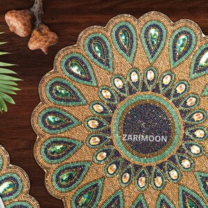 May include: A round beaded table mat with a peacock feather design. The mat is gold with green and blue accents. The center of the mat is a dark blue circle with the text "ZARIMOON" in white.
