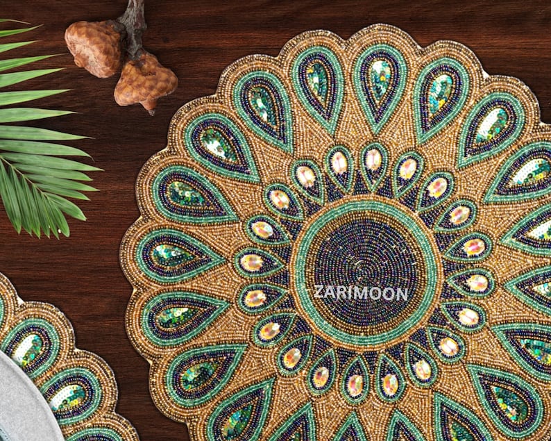 May include: A round beaded table mat with a peacock feather design. The mat is gold with green and blue accents. The center of the mat is a dark blue circle with the text "ZARIMOON" in white.