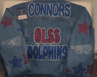 May include: A blue denim jacket with rhinestones spelling out "Connors" on the back, "OLSS" in red rhinestones, and "Dolphins" in blue rhinestones. The jacket also has red and blue star designs on the sleeves.