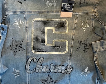 May include: A blue denim jacket featuring a large black and white rhinestone letter C and the word "Charm" in rhinestones. The jacket is adorned with several silver and black star-shaped rhinestones.
