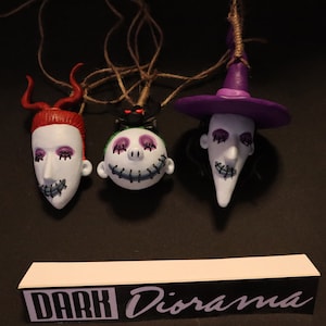Pode incluir: Três ornamentos de personagens de O Estranho Mundo de Jack, cada um com um rosto e um esquema de cores diferente. Os personagens estão pendurados em um barbante. Os ornamentos são brancos com detalhes em roxo, verde e vermelho. O texto "DARK Diorama" é visível em uma placa preta e branca na parte inferior da imagem.