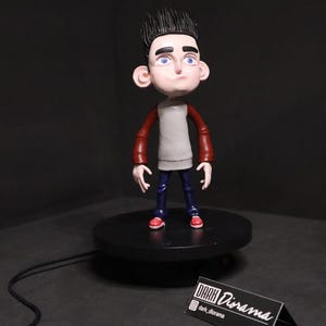 Puede incluir: Una figura de arcilla hecha a mano de un personaje de dibujos animados que lleva un suéter rojo y blanco, jeans azules y zapatos rojos. La figura está de pie sobre una base giratoria negra. La base tiene un cable negro conectado a ella. Un pequeño letrero con el texto "DARK Diorama" está en primer plano.