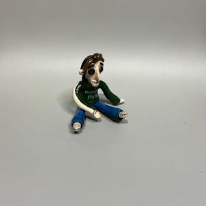 Può includere: Una piccola statuetta fatta a mano che raffigura una persona con capelli castani e occhi a bottone neri. Il personaggio indossa una maglia a maniche lunghe verde scuro con la scritta MICHIGAN STATE stampata in bianco, pantaloni blu e scarpe bianche.
