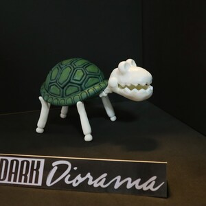 May include: A white skeletal turtle figurine with a green shell. The turtle is standing on a black surface with a sign that reads "DARK Diorama".