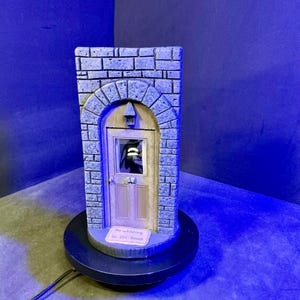 May include: A miniature decorative house facade with a stone brick exterior and arched doorway. The door is light brown, and the text "No whistling in the house" is visible on a small sign. The house sits on a black circular base.