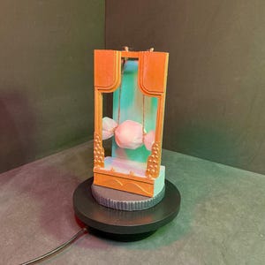 May include: A decorative, orange-colored, rectangular frame with a teal and white background. A pink, round object hangs from the top of the frame. The frame sits on a black, circular base.