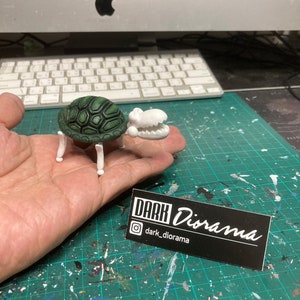 May include: A green and white turtle figurine with a white alligator head. The figurine is sitting on a person's hand. The background is a green and white checkered surface. The image also includes a black card with white text that says "DARK Diorama" and "@dark_diorama".