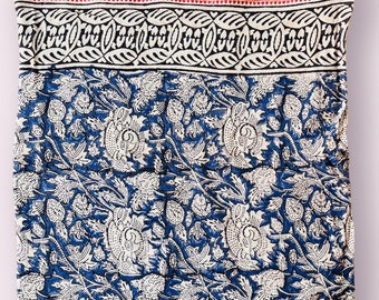 May include: A folded textile with a blue and white floral design. It has a border with black and white leaf patterns and a striped top section. The fabric is likely cotton or a similar material, suitable for a throw or a similar item.