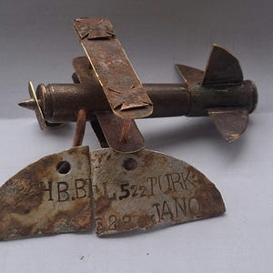 May include: A metal airplane sculpture crafted from a spent shell casing, featuring a propeller, wings with cross emblems, and a tail fin. Two weathered, semi-circular metal tags hang from the body, engraved with the text HB.BLL 522 TURK 2 JANO.
