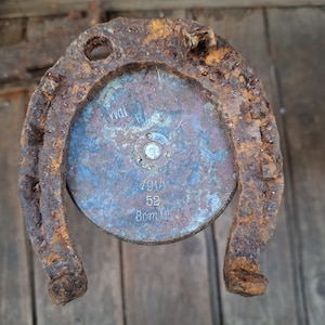 Puede incluir: Una herradura oxidada con un disco metálico redondo en el centro. El disco tiene la inscripción "W.D.I.R.M.G. 1914 52 8cm.Mb."