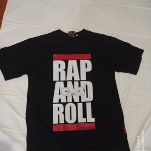 Peut inclure: T-shirt noir avec un motif blanc qui dit "RAP AND ROLL" avec une barre rouge derrière le texte. Le motif comprend également une paire de mains qui roulent une cigarette. Le texte "baccababba.co" est en bas du motif.