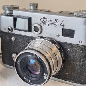 May include: Vintage silver and black FED 4 camera with a lens. The camera has a leather strap and the text "FED 4" is visible on the top. The lens has measurement markings and the number "032422".