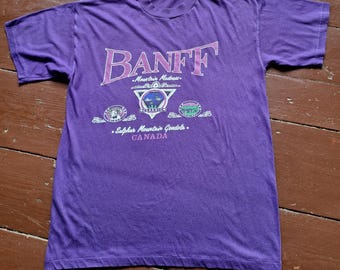 Könnte beinhalten: Lilafarbenes T-Shirt mit einem weißen Aufdruck des Wortes "BANFF" in rosa Schrift. Der Aufdruck enthält auch die Texte "Mountain Madness", "Sulphur Mountain Gondola" und "CANADA".