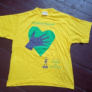 Könnte beinhalten: Gelbes T-Shirt mit einem grünen Herzen und einer Hand darin. Der Text auf dem Shirt lautet "Children's Promise The Millennium Appeal" und "Made possible by Marks & Spencer".