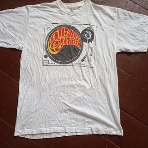 May include: White t-shirt with a graphic of a record player and the text "Orange Ethnic" in a yellow, orange, and red font.