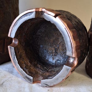 Puede incluir: Un cenicero de metal oxidado con un borde de cobre y un revestimiento de plata. El cenicero es redondo y tiene una abertura ancha.